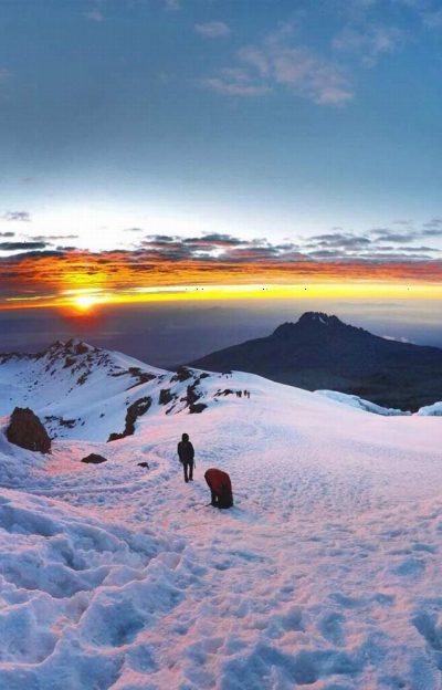 Climbers strung out across the snowy summit plateau at dawn, with Mawenzi in silhouette.