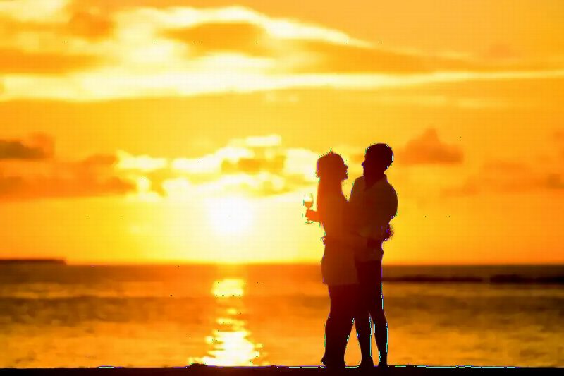 A couple embracing in silhouette against an orange sunset over water.