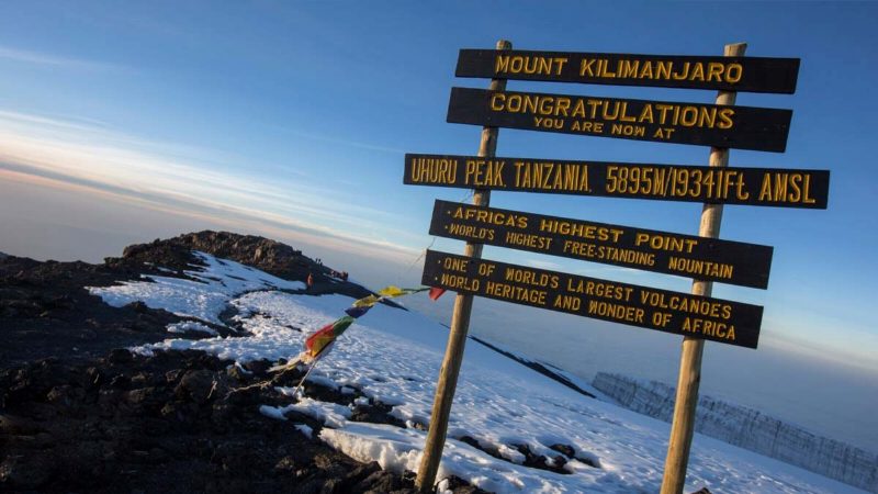 The Uhuru Peak summit sign at first light, prayer flags on one post and a glacier wall behind.