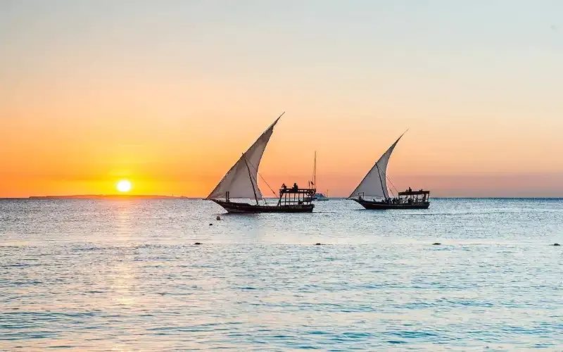 Two dhows sailing across calm water at sunset.