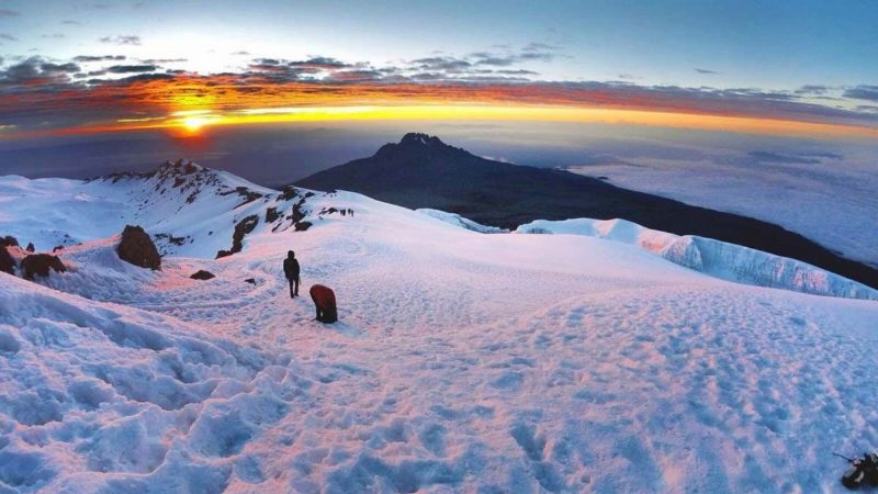 Two climbers crossing the snowy summit plateau at sunrise, with Mawenzi and orange cloud beyond.