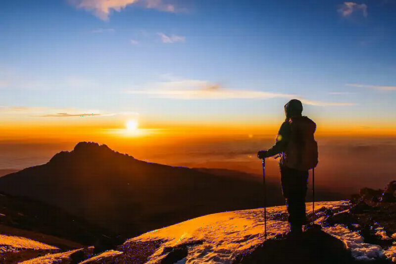 A climber silhouetted against a blazing sunrise high on the mountain, a jagged peak beyond.