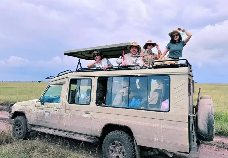 Three travellers standing up through the roof of a safari vehicle, laughing and waving.