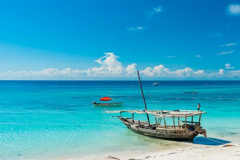 A wooden dhow anchored in clear turquoise water off a white beach.