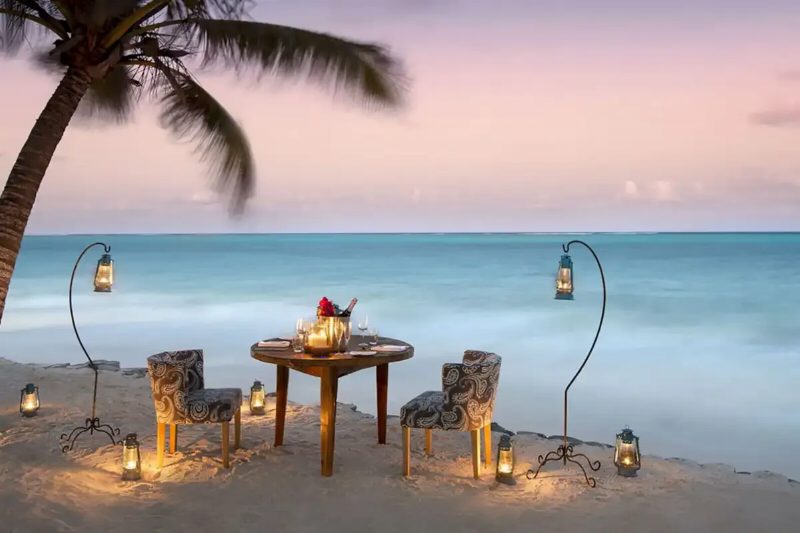 A table for two on the sand at dusk, with lanterns either side and the sea behind.