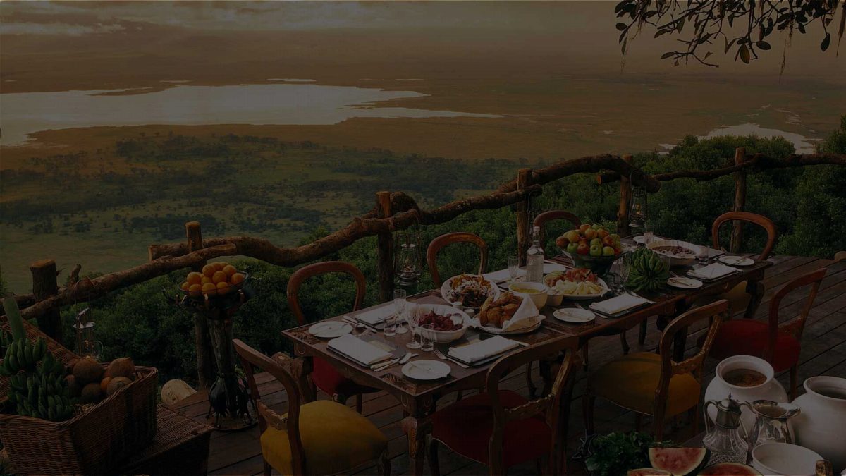 A dinner table on a lodge deck, lanterns along the railing, with the crater floor and soda lake below.