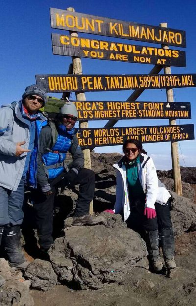 Three trekkers in down jackets beside the Uhuru Peak sign, snow and dark rock underfoot and clear blue sky behind.