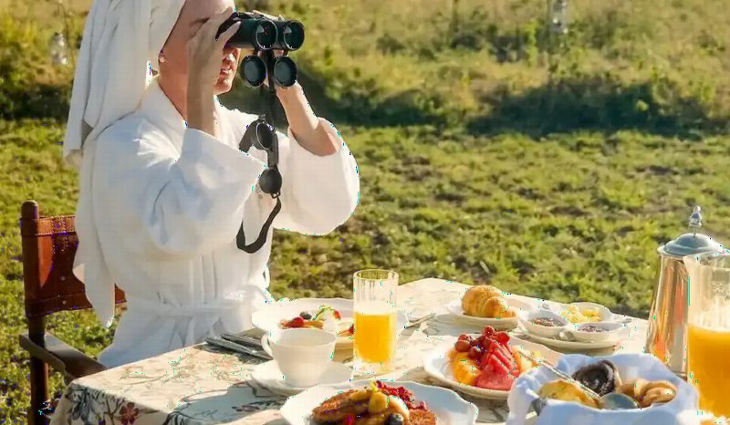 A guest in a white robe looking through binoculars over a breakfast table laid with fruit and juice.