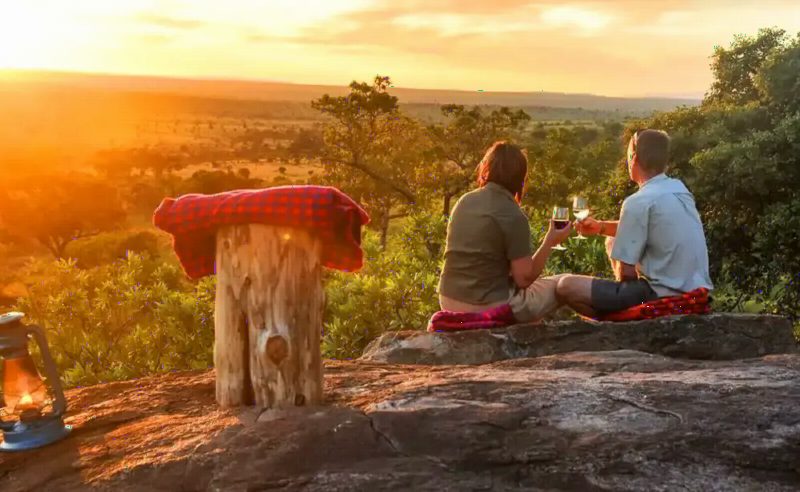 Two travellers sitting on a rock at sunset with drinks, a red blanket spread beside them.