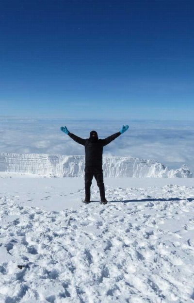 A climber on the summit plateau with both arms raised, a glacier wall behind and cloud to the horizon.