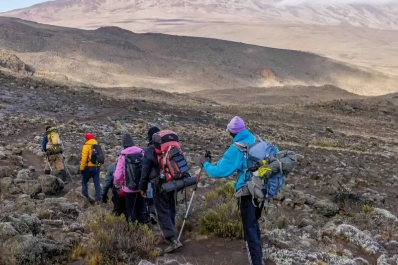 Trekkers crossing high, bare desert on the mountain, with a dusty ridge behind them.