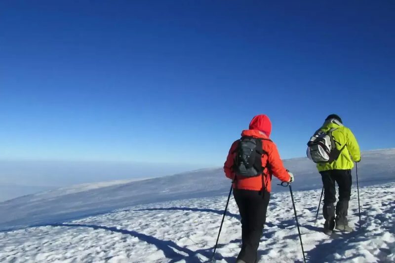 Two climbers in red and green jackets on a snowy slope above the clouds.