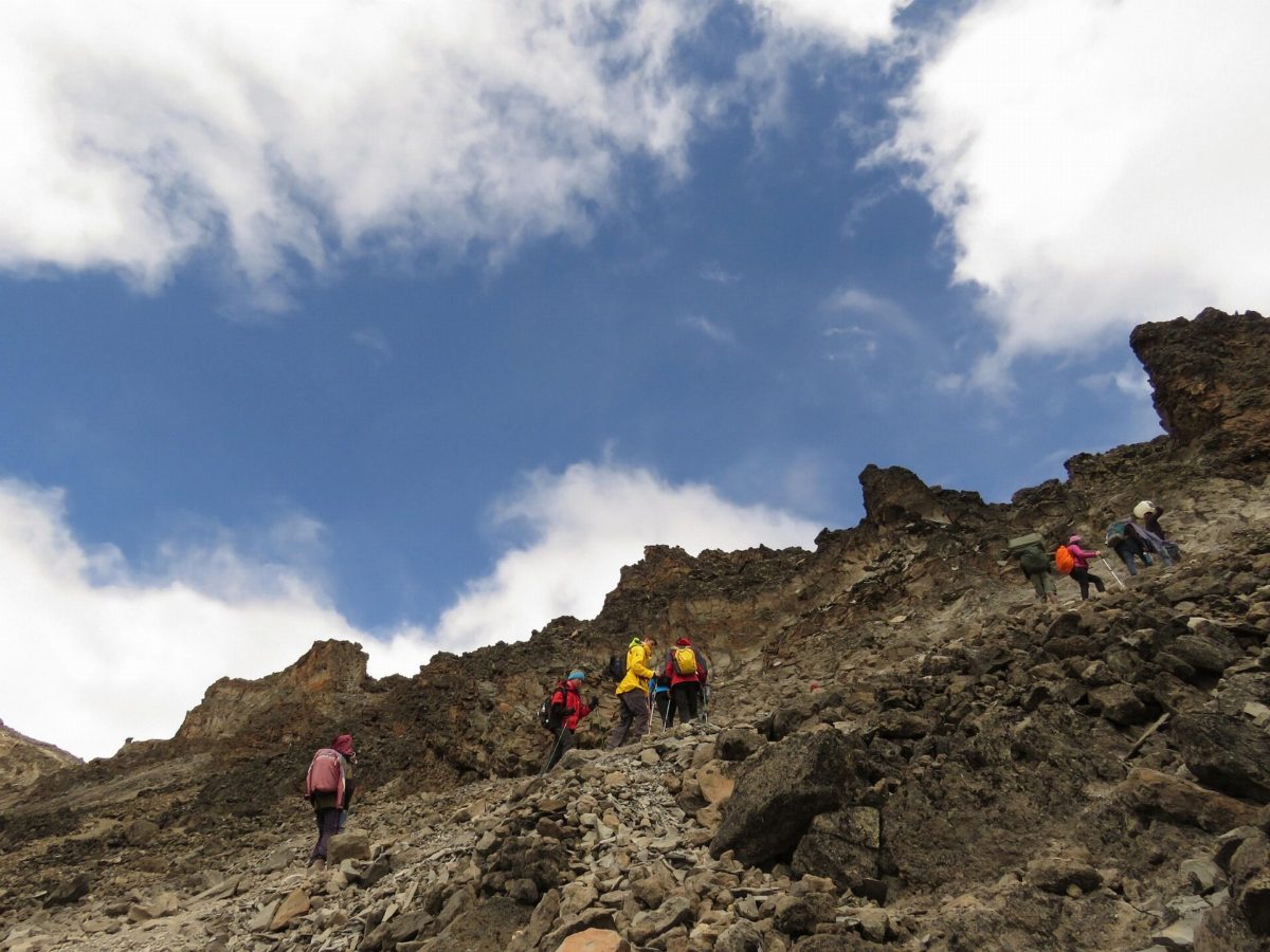 Climbers picking their way up a steep, rocky slope under a cloudy sky.