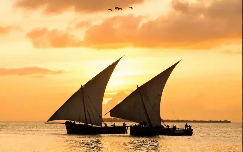 Two dhows with triangular sails silhouetted against an orange sunset sky.